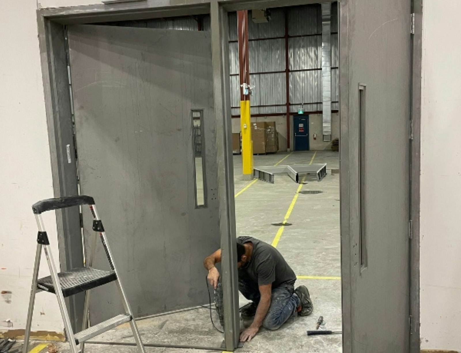 Door technician kneeling with a drill to fit a heavy steel door into its frame