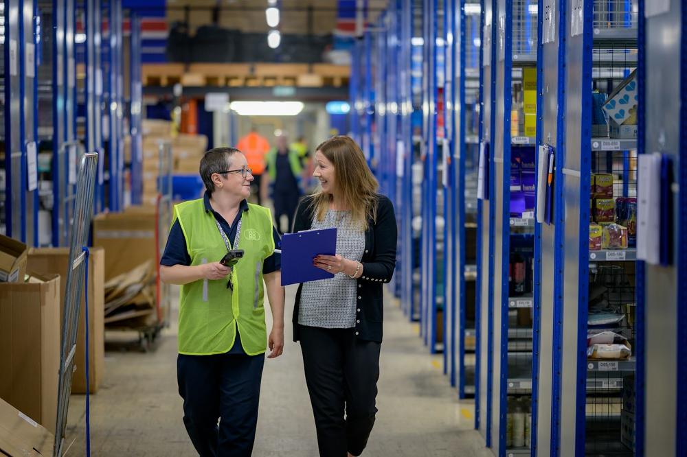Operations coordinator walking the warehouse floor with a clipboard, talking through the day's jobs